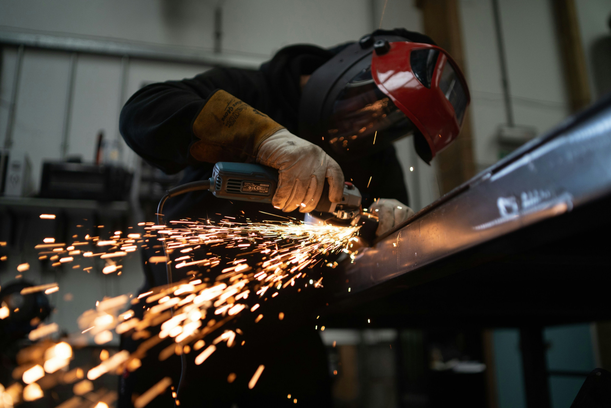 Worker in red welding helmet using angle grinder on metal beam with sparks flying