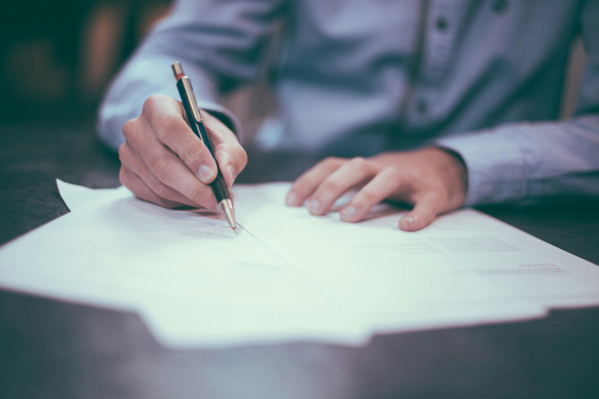 Person in light blue shirt signing a stack of formal documents on a dark table