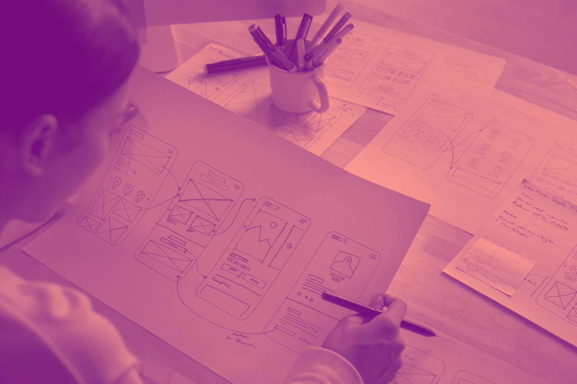shutterstock_2551665747 Overhead view of hands sketching app wireframes with purple-to-pink duotone treatment